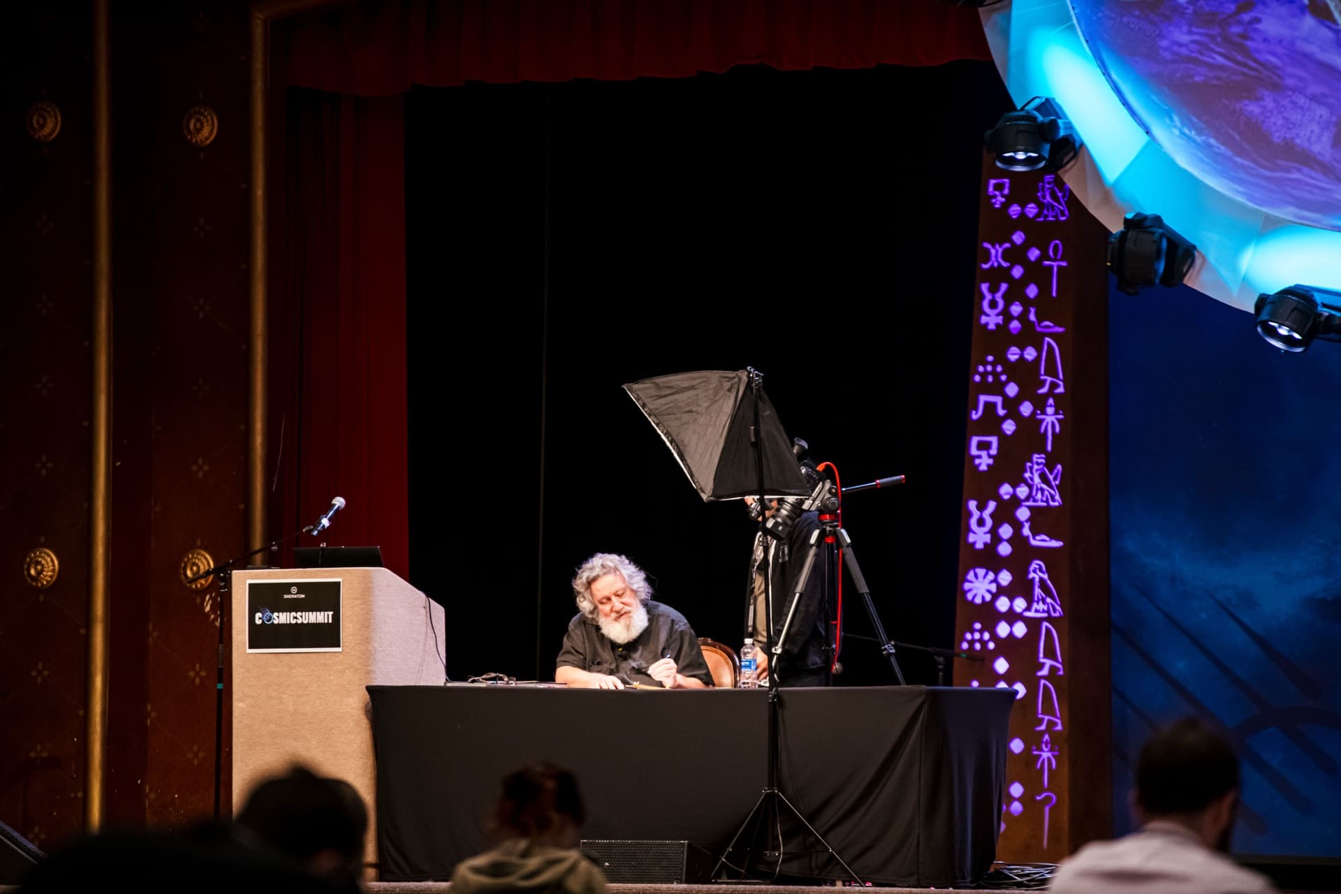 Cosmic Summit stage with a presenter at the desk and hieroglyph backdrop